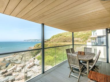 A covered balcony with wooden table and chairs overlooking the sea and rocky coastline at 17 Burgh Island Causeway in Bigbury-On-Sea