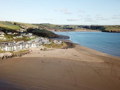 A coastal area with a sandy beach a parking lot buildings and green hills in the background at 17 Burgh Island Causeway in Bigbury-On-Sea