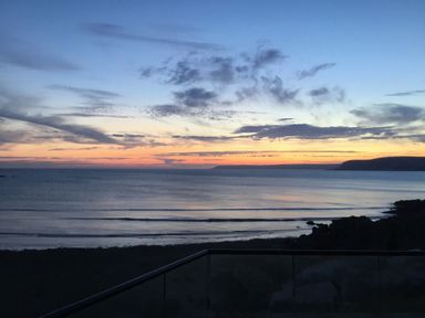 A sunset over the ocean with a beach and rocky shore viewed from behind a glass railing at 17 Burgh Island Causeway in Bigbury-On-Sea