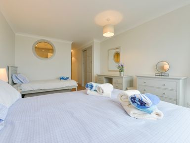 A bedroom with two beds, a dresser with a round mirror, a desk with a mirror and flowers, and towels on the larger bed at 17 Burgh Island Causeway in Bigbury-On-Sea
