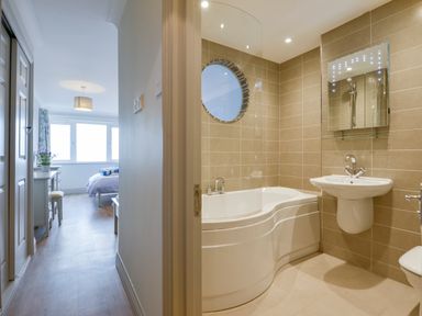 A bathroom with a curved bathtub sink and mirror next to a bedroom with a bed desk and window at 17 Burgh Island Causeway in Bigbury-On-Sea