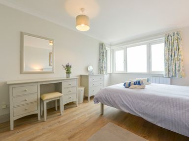 A bedroom with a bed towels on it a dressing table with a chair a mirror a chest of drawers and a window with curtains at 17 Burgh Island Causeway in Bigbury-On-Sea