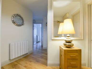A hallway with wooden flooring a wooden cabinet with a lamp and a mirror above and a wall clock on the left at 17 Burgh Island Causeway in Bigbury-On-Sea