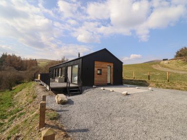 A black and wooden rectangular building on a gravel area with a deck and railing in a rural setting at The Sheep Shed in Llanrhaeadr-Ym-Mochnant