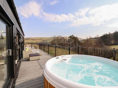 A deck with a hot tub sofa and table overlooking hills and trees at The Sheep Shed in Llanrhaeadr-Ym-Mochnant