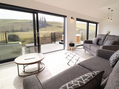 A living room with gray sofas, patterned cushions, a wood table, a small black stool, and glass sliding doors opening to a deck with a hot tub and a scenic countryside view at The Sheep Shed in Llanrhaeadr-Ym-Mochnant