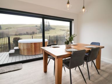A dining area with a wooden table set for four and a view of a hot tub outside through large glass doors at The Sheep Shed in Llanrhaeadr-Ym-Mochnant