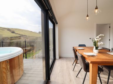 A dining area with a wooden table set with plates and chairs next to large glass doors opening to a balcony with a hot tub at The Sheep Shed in Llanrhaeadr-Ym-Mochnant