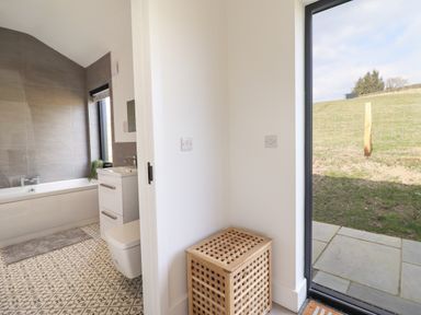 A bathroom with a bathtub toilet and sink next to a laundry basket and glass door showing outdoor grass at The Sheep Shed in Llanrhaeadr-Ym-Mochnant