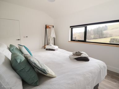 A bedroom with a white bedspread pillows and folded towels on the bed a window showing a countryside view and a clothes rack with robes at The Sheep Shed in Llanrhaeadr-Ym-Mochnant