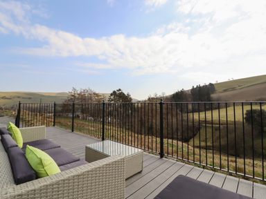 An outdoor deck with wicker sofa seating cushions and a glass coffee table overlooking a grassy hillside at The Sheep Shed in Llanrhaeadr-Ym-Mochnant