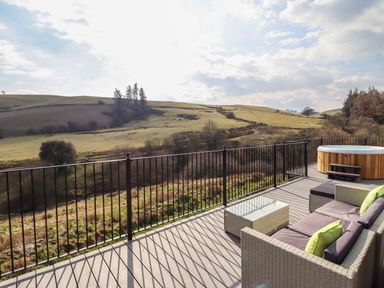 An outdoor deck with patio furniture a glass table and a hot tub overlooking grassy hills and trees at The Sheep Shed in Llanrhaeadr-Ym-Mochnant