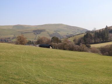A grassy hillside with trees and buildings in the valley and hills in the background at The Sheep Shed in Llanrhaeadr-Ym-Mochnant