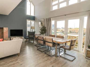 A dining area with a wooden table and six chairs near large glass doors and a living room with a sofa a TV and a tall window at The Deck House in Beadnell
