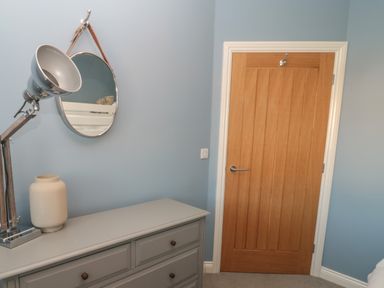 A room corner with a wooden door a gray dresser with a lamp and a round mirror hanging on the blue wall at The Deck House in Beadnell