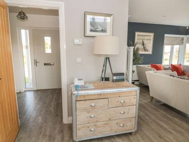 A hallway with a wooden chest of drawers and a lamp next to a living room with sofas and red cushions at The Deck House in Beadnell