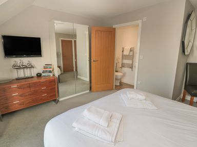 A bedroom with a bed with folded towels a wooden dresser with decorative items a wall-mounted tv a mirrored wardrobe an open door to a bathroom and a chair at The Deck House in Beadnell