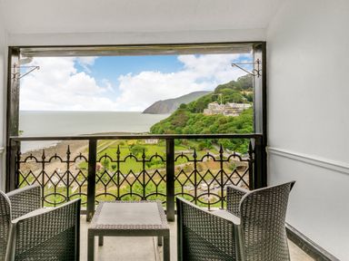 A balcony with two chairs and a table overlooking a coastal landscape with greenery and buildings at Clooneavin Apartment 4 in Lynmouth