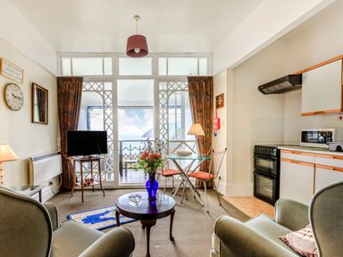 A living room with armchairs a small round table and a vase of flowers a TV on a stand and a glass dining table with chairs next to a kitchen area with appliances and cabinets at Clooneavin Apartment 4 in Lynmouth