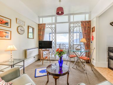 A living room with a glass dining table and metal chairs a television on a wooden stand a round wooden coffee table with flowers in a blue vase and a glass side table with a lamp at Clooneavin Apartment 4 in Lynmouth