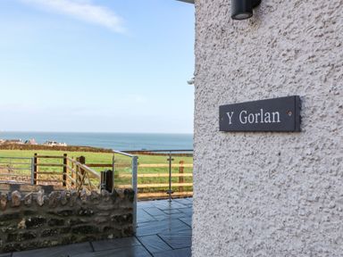 A view of a stone wall and outdoor area with a sign reading Y Gorlan and a grassy field and ocean in the background at Y Gorlan in Moelfre