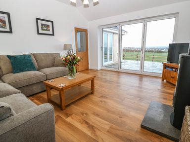 A living room with two sofas a wooden coffee table with flowers large sliding glass doors and a television at Y Gorlan in Moelfre