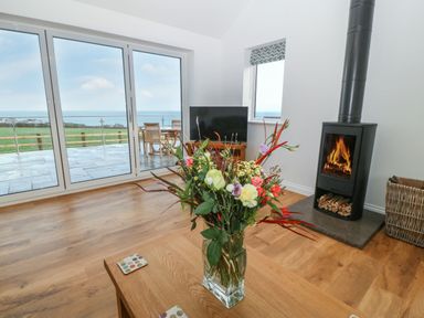 A living room with a vase of flowers on a wooden table a wood burning stove a television and sliding glass doors leading to a patio at Y Gorlan in Moelfre
