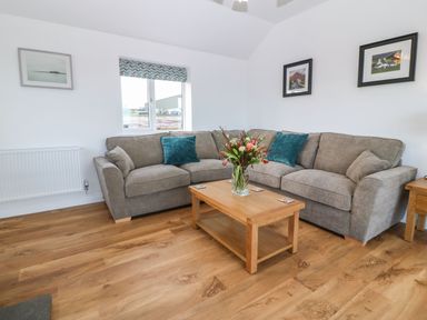 A sitting room with a grey corner sofa blue cushions wooden coffee table with flowers and framed pictures on white walls at Y Gorlan in Moelfre