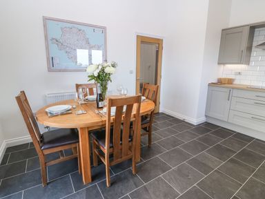 A dining area with a wooden table and four chairs set with plates and glasses next to a kitchen with grey cabinets and tiled floor at Y Gorlan in Moelfre