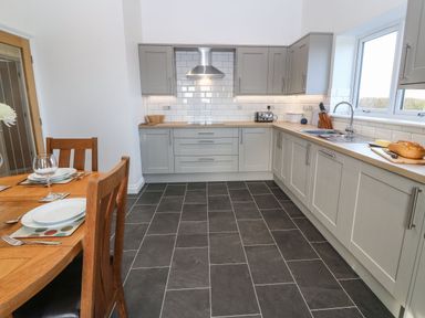 A kitchen with grey cabinets a wooden dining table set with plates and glasses and a window above the sink at Y Gorlan in Moelfre