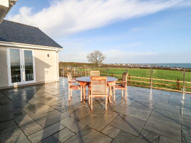 A patio with a round wooden table and four wooden chairs overlooking a grassy field and distant houses at Y Gorlan in Moelfre