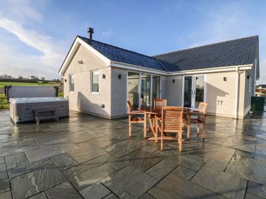 An outdoor patio with a round wooden table and four chairs next to a house with sliding glass doors and a hot tub at Y Gorlan in Moelfre
