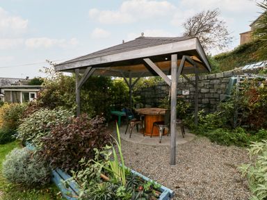 A wooden gazebo with a round table and metal chairs on a gravel surface surrounded by plants and a stone wall at Gerlan in Harlech