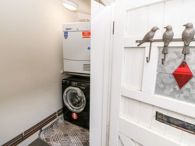 A laundry room with a washing machine and a dryer stacked in a corner and a white door with bird-shaped hooks and stained glass at Gerlan in Harlech