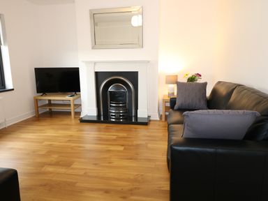 A living room with a black leather sofa a fireplace a TV on a wooden stand and a side table with a lamp and flowers at Gatehouse in Moville County Donegal