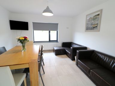 A living room with a wooden dining table and chairs a vase of flowers a wall-mounted television two leather sofas a painting and a window with a blind at Gatehouse in Moville County Donegal