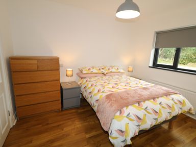 A bedroom with a bed with patterned bedding two bedside tables with lamps a wooden dresser a window with a gray blind and wooden floor at Gatehouse in Moville County Donegal