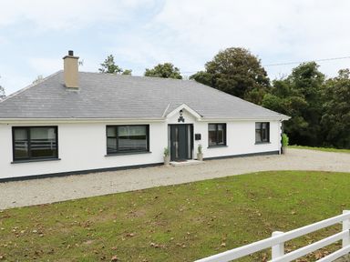 A single story house with a gray roof white walls and black window frames surrounded by grass and trees at Gatehouse in Moville County Donegal