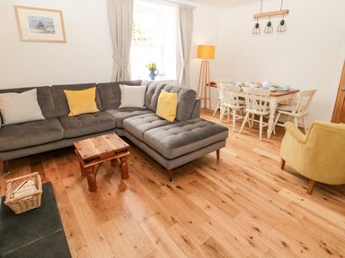 A living room with a gray sectional sofa yellow and white cushions a wooden table a basket of wood a dining table with chairs and a yellow armchair at Curlew Cottage 2 Fenkle Street Holy Island