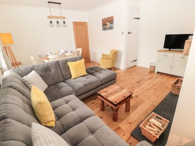 A living room with a gray sectional sofa yellow cushions wooden coffee table dining table with white chairs and a television on a cabinet at Curlew Cottage 2 Fenkle Street Holy Island
