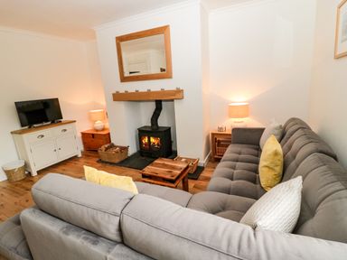 A living room with a gray sectional sofa yellow and patterned pillows two side tables with lamps a wood stove with fire a mirror above and a TV on a white cabinet at Curlew Cottage 2 Fenkle Street Holy Island