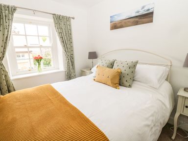 A bedroom with a bed with white sheets green and yellow pillows and a yellow blanket next to a window with green curtains and flowers at Curlew Cottage 2 Fenkle Street Holy Island