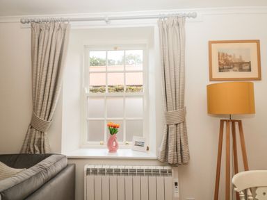 A window with beige curtains a pink vase with orange flowers a framed photo a floor lamp a gray couch and a framed picture on the wall at Curlew Cottage 2 Fenkle Street Holy Island