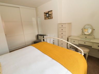 A bedroom with a bed covered with white and yellow linens a chair a tall dresser a dressing table with a mirror and a wardrobe with sliding doors at Curlew Cottage 2 Fenkle Street Holy Island