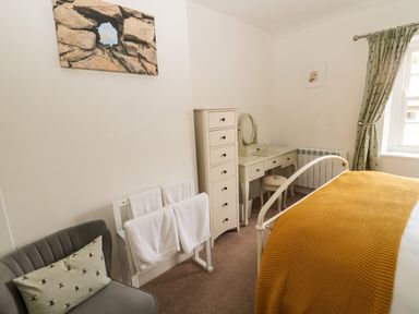 A bedroom with a grey chair a white towel rack a dresser a vanity table and a bed with a yellow blanket at Curlew Cottage 2 Fenkle Street Holy Island