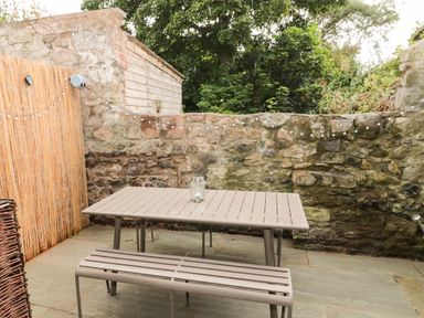 An outdoor patio with a table and bench a stone wall and string lights in the background at Curlew Cottage 2 Fenkle Street Holy Island