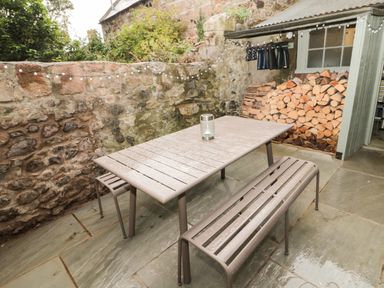 An outdoor patio area with a stone wall a metal table and benches a stack of firewood and boots hanging at Curlew Cottage 2 Fenkle Street Holy Island
