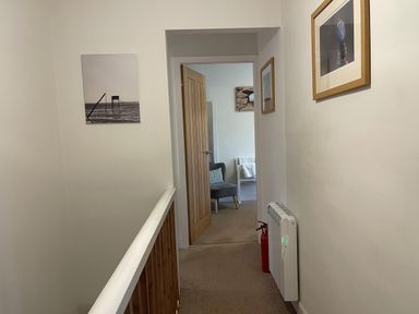 A hallway with cream walls and carpet leading to a room with a gray chair and light wooden door at Curlew Cottage 2 Fenkle Street Holy Island