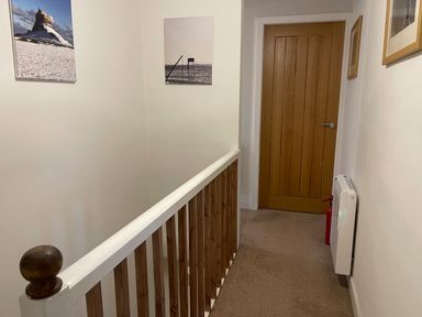 A hallway with a wooden banister a closed wooden door a fire extinguisher and framed pictures on the walls at Curlew Cottage 2 Fenkle Street Holy Island