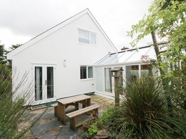 An outdoor patio area with a wooden picnic table and bench surrounded by plants and a white house with glass conservatory at Alice Cottage in Wheal Kitty near St Agnes
